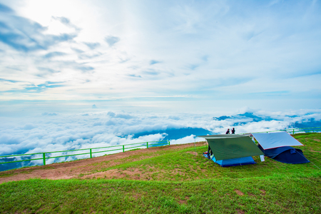 Man in orange jacket looking at view of mountain landscape with waves of fog and cloudy sky. Blue yellow camping tents on mountain peaks at Phu Tub Berg Phetchabun province Thailandの写真素材