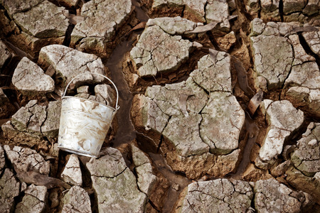 empty watering bucket on dried, cracked earth without water leftの写真素材
