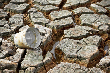 empty watering bucket on dried, cracked earth without water leftの写真素材