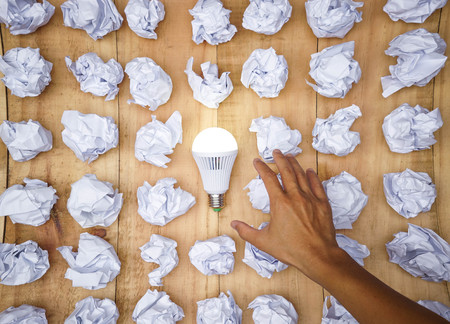 Hand trying to catch a turned on LED light bulb surrounded by crumpled paper ball / Business with new bright ideas and innovation conceptの写真素材