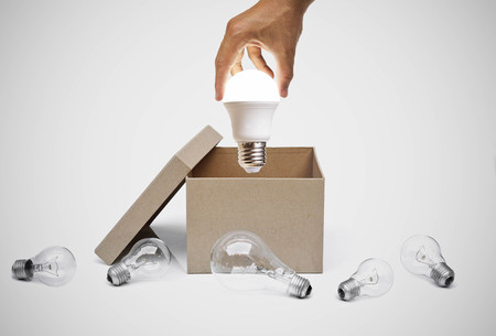Hand of a businessman holding a turned on LED light bulb coming out from a brown paper box surrounded by old incandescent light bulbs / Business with new idea and innovation conceptの写真素材