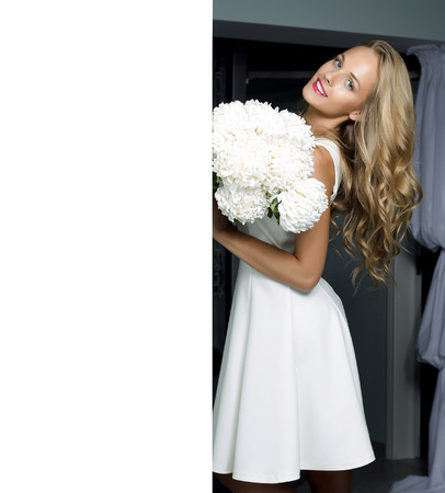 Awesome beautiful happy smiling woman with curly hair in white elegant dress poses holding flowers on stylish interior backgroundの写真素材