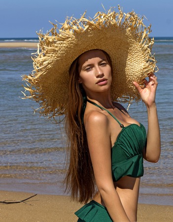 Portrait of woman in hat on beach. Beautiful gorgeous girl wearing green bikini poses near seaの写真素材