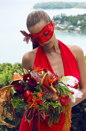 Beautiful woman in carnival mask with bouquet flowers wearing red dress poses on tropical sea view. Fashion masquerade look. Happy New Year festival. Phuket, Thailandの写真素材