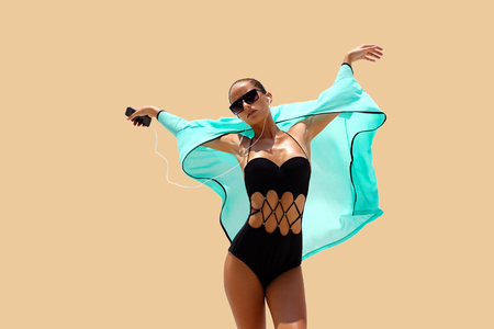 Dancing woman with headphones in black swimsuit and sunglasses poses on color background. High fashion look.の写真素材