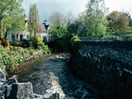 A quaint house sits by a gently flowing river, framed by vibrant greenery and foliage. The morning light illuminates the scene, highlighting the stone wall that borders the peaceful waterway. Trees sway gently in the breeze, creating a serene atmosphere in this picturesque location.の写真素材