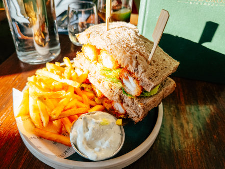 A hearty fish sandwich stacked high with fresh ingredients is paired with crispy fries and a small bowl of dipping sauce on a wooden table, illuminated by natural light.の写真素材