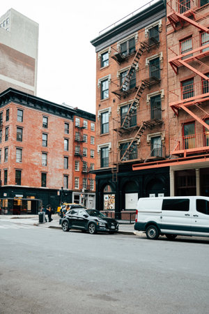 Two brick buildings stand at the corner of a bustling urban street, displaying fire escapes and parked cars under overcast skies. The atmosphere is lively yet tranquil, hinting at a vibrant community.の写真素材