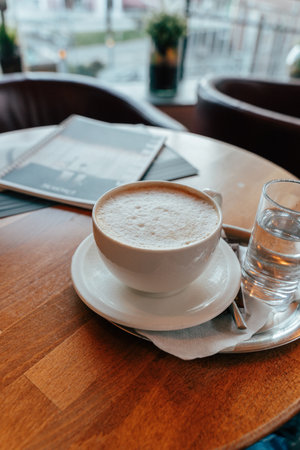 A steaming cup of coffee sits on a white saucer alongside a clear glass of water. The wooden table adds warmth to the inviting atmosphere of the cafe, with a menu in the background.の写真素材