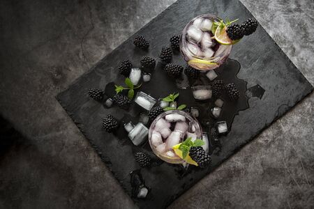 Seasonal soft drinks.Thirst in hot summer time. Two glasses of ice ,water ,lime and mulberry berries with mint on a dark background .Keto diet,soft drinks and alcoholic beverages. Fruit cocktailの写真素材