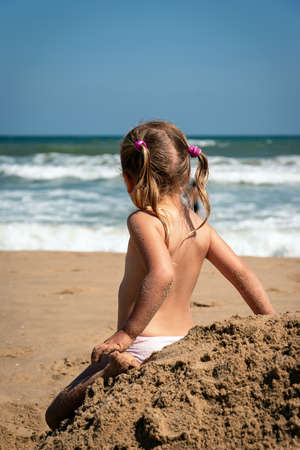 Baby girl in summertime rear view. Girl child relax alone at seaside. Preschool blonde looking at the ocean waves. Adorable little kid sitting back at pile of sand. Pensive child at seashore scenery.の写真素材