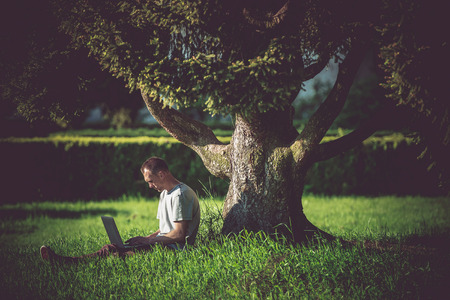 Internet Break Under the Tree. Men Enjoying Wireless Internet Connectivity. WiFi Connection in the Garden. Caucasian Men Working Remotely on His Laptop Computer.の写真素材