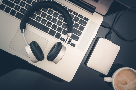 Freelance Worker Mobile Desktop Workstation. Laptop Computer, Headphones, Small Portable Hard Drive and Cup of Fresh Cappuccino Coffee on Black Glassy Desk Surface. Freedom of Freelance Work Concept.の写真素材