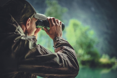 Caucasian Hunter in Masking Camouflage Uniform with Binoculars. Hunter Spotting Game. Poacher or...