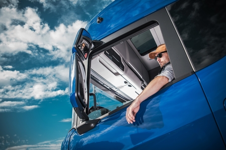 Heavy Load Cargo Delivery. Caucasian Truck Driver Inside Semi Tractor Cabin and the Blue Sky. Transportation Industry.の写真素材