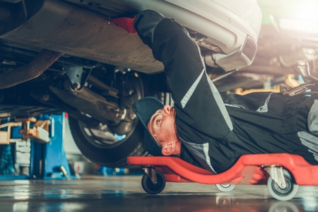 Caucasian Car Mechanic in His 30s Under the Car on the Mechanics Creeper Trying To Fix Modern Vehicle Exhaust System.の写真素材