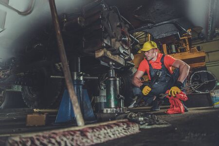 Heavy Equipment Repair Shop. Caucasian Professional Mechanic Fixing Excavator Undercarriage....