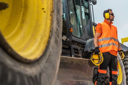Road Construction Business. Caucasian Heavy Machinery Operator in His 30s with Noise Reduction Headphones and Hat Protection Helmet in a Hand.の写真素材