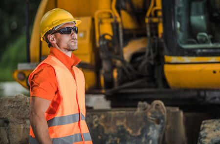 Construction Worker on Duty. Caucasian Men in His 30s Wearing Safety Accessories. Building Machinery in a Background. Industrial Zone.の写真素材