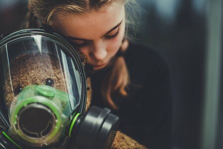 Sad Caucasian Girl Hugging Her Teddy Bear Toy Who Wearing Biochemical Gas Mask.の写真素材