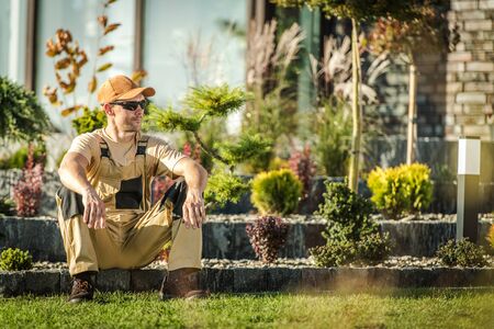 Mid 30s Male Gardener Sitting Down And Relaxing On Stone Edging In Residential Backard Area. の写真素材