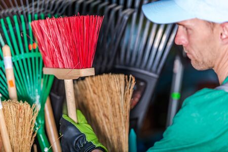 Close Up Of Caucasian Male Groundskeeper Choosing Small Broom In Storage Shed.
