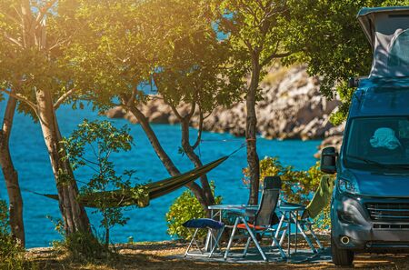 Scenic Outdoor Camping Spot. Motorhome, Garden Chairs and Hammock Between Trees. Sea Shore Scenery. Vacation Time in Camper Van.の写真素材