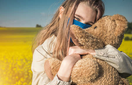 Portrait of Happy Caucasian Teenage Girl in Face Mask Hugging Her Teddy Bear During Pandemic. Virus Outbreak Theme.の写真素材