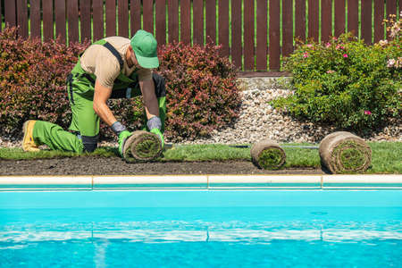 New Grass Turfs Installation Along Side of Garden Outdoor Swimming Pool. Caucasian Gardener in His 40s Assembly Grass From Rolls. Landscaping Industry.の写真素材