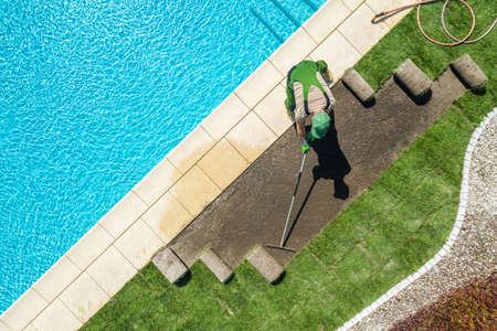 Caucasian Professional Landscaper Installing Brand New Grass Turfs Around Residential Swimming Pool. Raking Soil Before Installation. Gardening and Landscaping Industry Theme.の写真素材