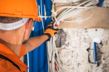 Caucasian Construction Contractor in His 40s Wearing Hard Hat Looking Inside Electrical Installation. Grabbing Electric Cables. Industrial Theme.の写真素材