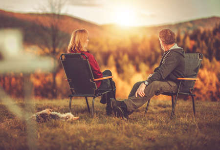 Caucasian Couple in Their 40s Seating in Front of Scenic Vista Enjoying Their Life and the Nature. Australian Silky Terrier Next to Chairs. Colorful Fall Foliage.の写真素材