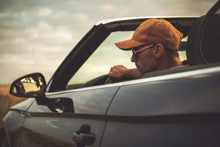 Caucasian Men in His 40s Wearing Baseball Cap Driving His Convertible Car with Roof Opened During Summer Road Trip. Automotive and Transportation Industry Theme. Cabrio Vehicle Drive.の写真素材