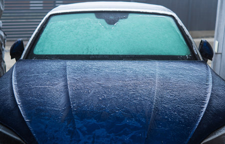 Modern Blue Body Car Covered in Frost During Freezing Cold Winter Morning. Front of the Vehicle. Seasonal Automotive Maintenance and Weather Elements.の写真素材