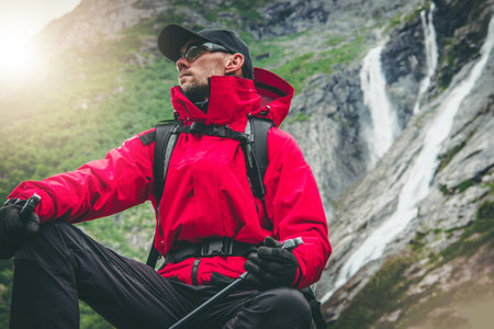 Outdoor Recreation. Active Caucasian Hiker Wearing Red Rainproof Jacket and Eyes Safety Glasses Seating Next to Scenic Alpine Waterfalls. Exploring the Trail.の写真素材