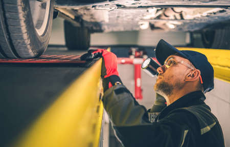 Caucasian Car Mechanic in His 40s with Flashlight Spotting Vehicle Suspension Problems. Automotive Service Job. Transportation Industry Theme.の写真素材