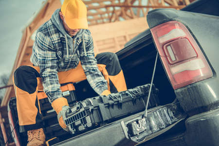 Caucasian Construction Contractor Worker Preparing His Tools on the Pickup Cargo Section. Wooden House Building in a Background. Industrial Theme.の写真素材