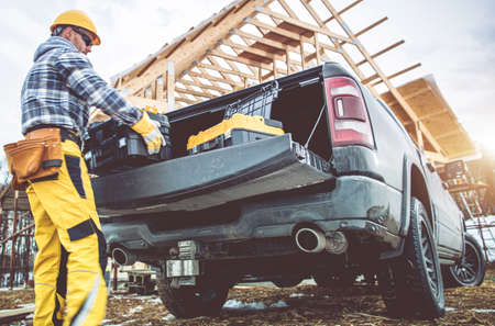 Caucasian Construction Contractor Worker in His 40s Taking Tools Boxes From Bed Section of His Pickup Truck. Newly Constructed Building in Background.の写真素材
