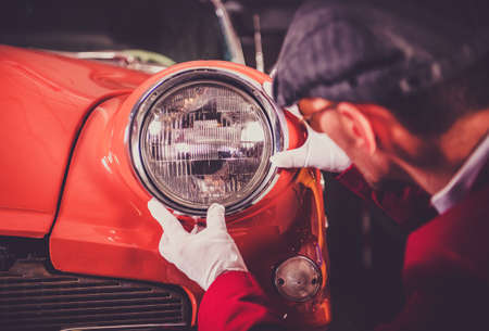 Caucasian Hobbyist in White Gloves Restoring Dark Orange Reddish Body Vintage Car. Front Headlamp Check. Classic Cars and Automotive Industry Theme.の写真素材