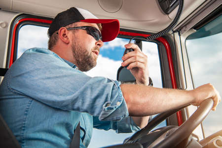 Caucasian Semi Truck Driver in His 30s Taking Conversation Using CB Radio Inside His Truck Cabin.