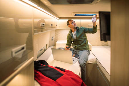 Caucasian Tourist in His 40s Seating in Front of His Laptop Computer Inside Camper Van Motorhome Opening Roof Vent to Get Some More Fresh Air Into His RV Recreational Vehicle.の写真素材
