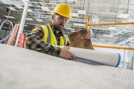 Construction Contractor Worker in His 30s Looking Inside Large Paper Building Blueprints....