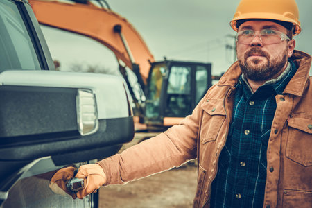 Ground Work Excavation Worker in Yellow Helmet and Safety Glasses Staying Next to Heavy Equipment in...