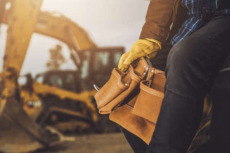 Professional Construction Worker with Tools Belt in His Hands. Excavator and Other Heavy Machinery in the Background. Close Up Photo. Industrial Theme.の写真素材
