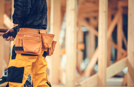 Residential Buildings Construction Worker with Tools Belt and Wooden House Skeleton Frame in a Background. Construction Industry Theme.の写真素材