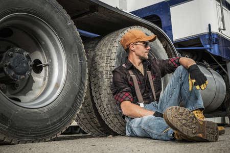 Semi Truck Driver Resting Next to His Truck Seating on a Ground. Heavy Duty Transportation Job Theme.の写真素材