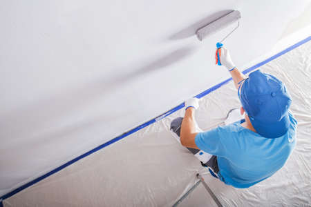 Male Contractor Wearing Blue T-Shirt and Blue Baseball Cap During Renovation Work Painting the Walls in a Room White with a Paint Roller.の写真素材