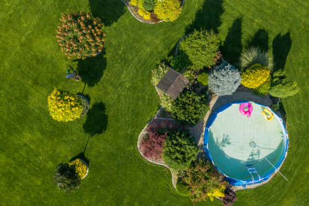 Beautifully Landscaped Garden at the Backyard with Swimming Pool and Perfectly Maintained Lawn. Professional Gardener at Work Taking Care of Plants. Hot Summer Day with Trees Casting Shadows on the Grass. Aerial View.の写真素材