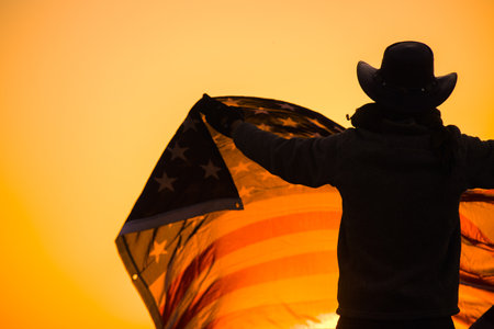 Silhouette of a Man Wearing a Western Style Hat Waving a US Flag against the Background of a Scenic Sunset. Celebration of the America's Independence Day on Fourth of July.の写真素材