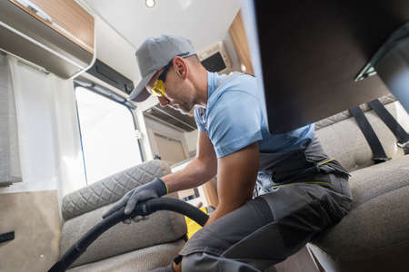 Male Worker in His 40s Cleaning the Sofa Bed Couch in a Camper Vehicle with a Vacuum Cleaner. Care and Maintenance of a Modern and Comfortable Camper Interior Adapted for Long-Term Travel.の写真素材